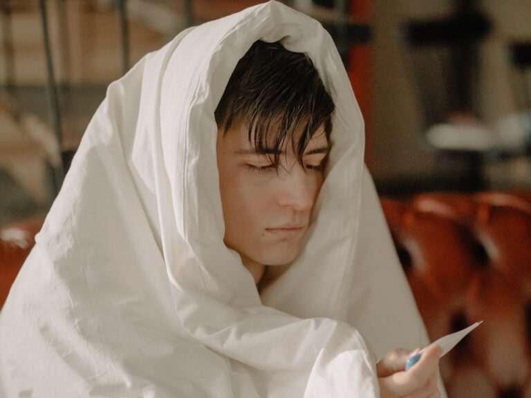 Young man covered in white blanket looking at thermometer