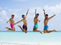 Four men in swim trunks jumping high in unison on beach